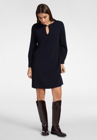Frau in einem knielangen schwarzen Langarmkleid mit einem Schlüsselloch-Ausschnitt und hohen glänzenden dunkelbraunen Lederstiefeln.