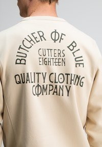 Beige sweatshirt met opvallende zwarte tekst: "BUTCHER OF BLUE," "CUTTERS EIGHTEEN," en "QUALITY CLOTHING COMPANY" op de achterkant. Zachte stof, ronde hals.