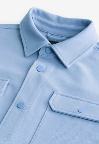 Camisa abotonada de color azul claro, texturizada, con collar de punta, dos bolsillos en el pecho y botones en un tono azul claro contrastante.