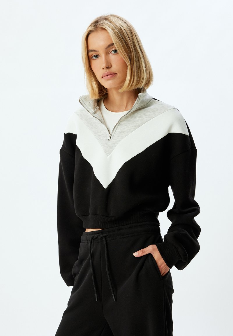 Sudadera negra cropped con cuello de medio zip, que presenta un diseño en chevrón gris y blanco en el pecho. Tejido suave, corte relajado.