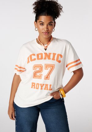 Femme aux cheveux bouclés portant une chemise blanche avec le texte orange « ICONIC 27 ROYAL », un jean bleu et des bracelets, posant avec une main sur la hanche.