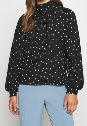 Schwarze Langarmbluse mit weißen Polka-Dots und einer gebundenen Schleife am Ausschnitt, kombiniert mit hellblauen Hosen.