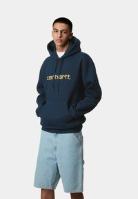 Sudadera con capucha azul marino y logo amarillo de "carhartt", con bolsillo canguro y puños acanalados, combinada con pantalones cortos de mezclilla azul claro.