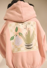 Różowa bluza z kapturem z dużą ilustracją z tyłu, przedstawiającą kwiaty, gruszkę i zielone liście na kraciastym tle. Gładka tekstura materiału.