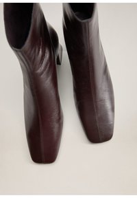 Botas de cuero marrón oscuro hasta el tobillo con puntera cuadrada y tacones bajos, mostradas desde un ángulo superior sobre un fondo neutro.