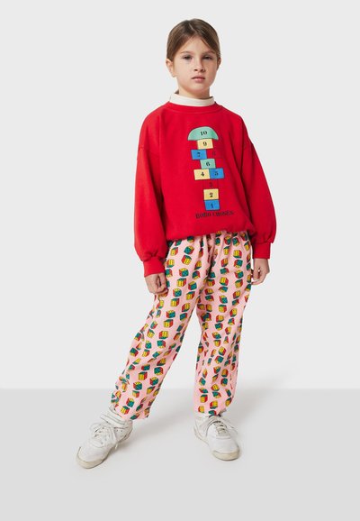 Felpa rossa con un grafiche di campana; pantaloni rosa con motivi a blocchi colorati. Modello indossa sneakers bianche. Look casual e comodo.