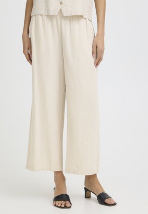 Žena má na sebe voľné béžové široké nohavice, k nim čierne otvorené mule na podpätku, stojaca proti jednoduchému svetlému pozadiu.