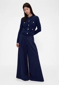 Cardigan de malha azul-marinho com grandes botões dourados e bolsos frontais, combinado com calças largas azul-marinho com riscas verticais.