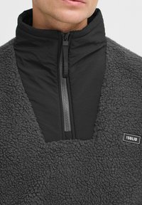 Gray sherpa pullover with a high collar, featuring a zip front. The top layer is smooth, while the lower portion has a fluffy texture. Black logo detail.