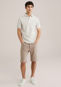 Gai bēšas krāsas polo krekls ar īsām piedurknēm, kopā ar bēšām šortiem. Apģērbs ietver baltas sporta kurpes un ir izgatavots no teksturēta auduma.