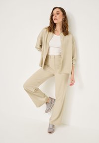 Chemise beige oversize à boutons, débardeur blanc, pantalon beige ajusté et baskets grises avec des accents bruns. Texture lisse et coupe décontractée.