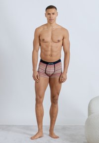 Homme athlétique debout, pieds nus, portant un shorty rayé avec une ceinture marine, sur un fond clair uni.