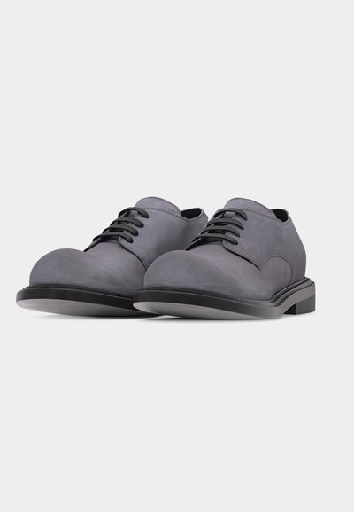 Paire de chaussures habillées en daim gris à lacets, bouts ronds et semelles noires sur un fond gris clair uni.