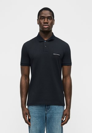 Vyras dėvi juodą trumpomis rankovėmis polo marškinėlį su Armani Exchange logotipu ir šviesiai mėlynas džinsus, stovintis prieš paprastą foną.