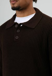 Man draagt een donkerbruine geribbelde trui met kraag en drie knopen over een wit shirt met ronde hals, gefotografeerd vanaf net onder het gezicht en de schouders.