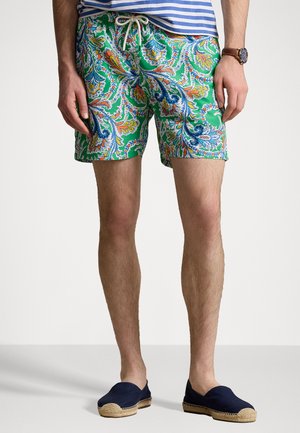 Grüne Badeshorts mit einem Paisleymuster, das orangefarbene und blaue Akzente aufweist, und einem Kordelzugbund. Getragen mit marineblauen Espadrilles.
