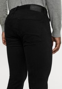 Černé džíny z denimu se rovného střihu, obsahující texturovanou tkaninu a decentní zadní kapsy. Na pase je přišitá kožená nášivka.