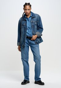 Jeansjacke mit einem verblassten blauen Finish, über einem passenden Denim-Hemd getragen, kombiniert mit hellblauen Jeans. Schwarzer Gürtel und klobige schwarze Schuhe.