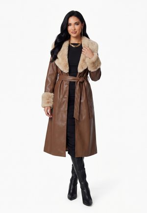 Donna che indossa un cappotto di pelle marrone con colletto e polsini in pelliccia beige, abito nero, gioielli in oro e stivali neri al ginocchio, in piedi su sfondo bianco.