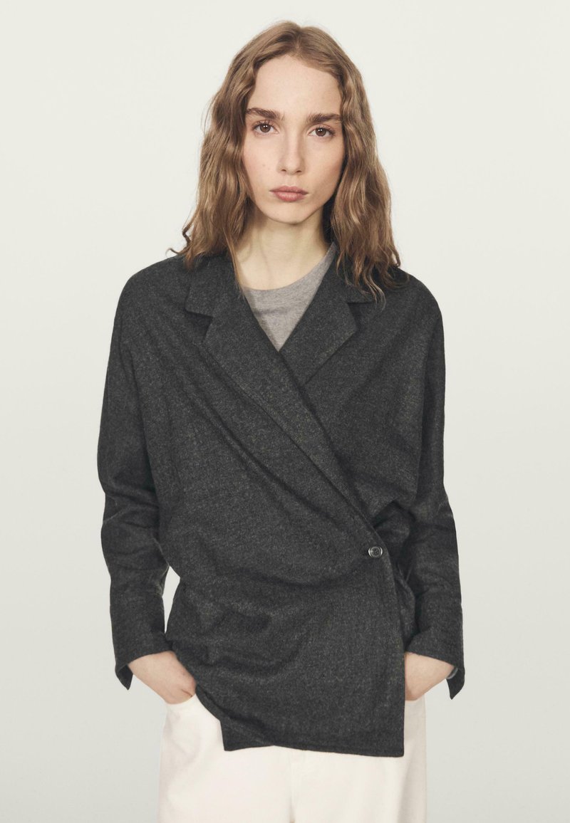 Houtskoolgrijze, gestructureerde wikkeljacket met een geknipte kraag, enkel knoopsluiting en lange mouwen, gelaagd over een grijze shirt.