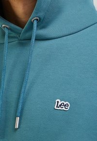 Sudadera turquesa con una textura suave, que cuenta con un bolsillo canguro, terminales de metal plateado en los cordones y un logo bordado de "Lee".
