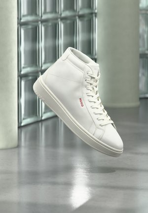 Weißer High-Top-Sneaker mit Schnürsenkeln und rotem "Hugo"-Logo an der Seite, schwebt über reflektierendem grauem Boden in moderner Innenumgebung.