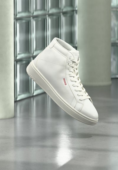 Baskets montantes blanches avec lacets et logo rouge "Hugo" sur le côté, flottant au-dessus d'un sol gris réfléchissant dans un cadre intérieur moderne.