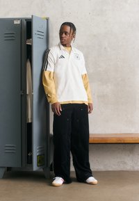 Polo atlético branco com riscas pretas, camada interior amarela e calças largas pretas. Apresenta sapatos brancos com detalhes vermelhos, ao lado de um armário cinza.