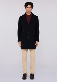 Cappotto in misto lana nero con taglio classico, rever a intaglio, tre bottoni e tasche laterali, abbinato a pantaloni chiari e scarpe scure.