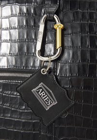 Schwarze Handtasche aus Krokodilleder mit silbernem Karabiner und einem schwarzen Tag mit der Aufschrift "ARIES" in Weiß.