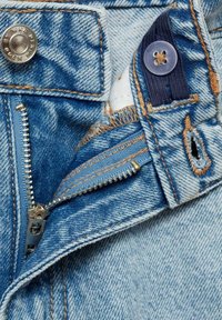 Denimstoff in hellblauer Farbe, mit einem Zipper mit Metallzähnen, einem Knopfverschluss mit graviertem Logo und kontrastierenden orangen Nähten.