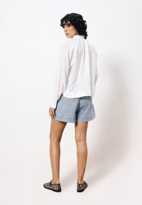 Weißes, strukturiertes Blusenoberteil mit langen Ärmeln; hellblaue Jeansshorts; schwarze und beige Slip-On-Schuhe mit Leopardenmuster; glatte Haut, sichtbare Tätowierungen.
