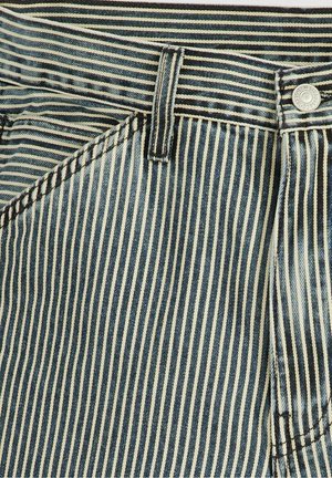 Nærbillede af blå og hvide vertikalt stribede denimstof med lomme, bæltestrop, syning og metal knap på linningen.