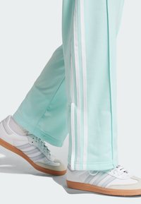 Mintgroene trainingsbroek met drie witte strepen langs de zijkanten, gecombineerd met witte sneakers met grijze accenten en een gumrubberen zool.