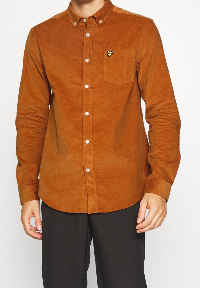 Camisa de botones de pana marrón con un bolsillo en el pecho y un pequeño logo. Mangas largas y dobladillo redondeado. Textura suave, ajuste estándar.