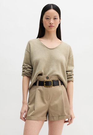 Vrouw met lang zwart haar, gekleed in een beige, lange mouw, gebreide trui en hoge taille kaki shorts met een zwarte riem.