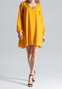 Gelbes V-Ausschnitt-Kleid mit langen, luftigen Ärmeln und fließender A-Linie, kombiniert mit schwarzen, geschlossenen High Heels vor einem grauen Hintergrund.