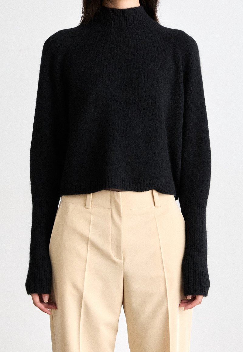 Femme portant un pull court noir à manches longues en maille côtelée, associé à un pantalon beige plissé taille haute.