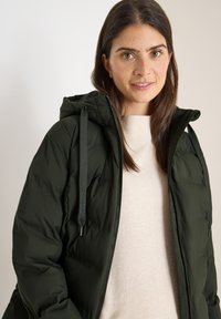 Giacca a vento verde scuro con cappuccio, dotata di corde regolabili e texture liscia. Indossata sopra un maglione beige chiaro.