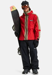 Man in een rode en beige jas en zwarte sneeuwbroek, die een zwart snowboard met een gele en rode punt vasthoudt. Hij draagt zwarte handschoenen, schoenen, een beanie en een skibril.