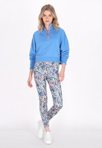 Kék cipzáras pulóver, magas gallérral, színes mintás leggings-szel, amelyeken sokszínű absztrakt formák láthatók, és fehér sportcipővel párosítva.