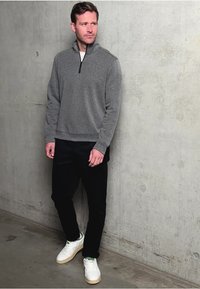 Muž v šedém svetru s čtvrtinovým zipem, černých kalhotách a bílých teniskách stojí proti betonové zdi a dívá se do strany.