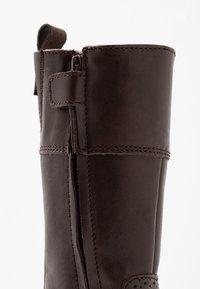 Brązowy skórzany but o gładkiej fakturze, z bocznym zamkiem błyskawicznym, wzmocnionym szyciem i pętelką u góry ułatwiającą zakładanie.