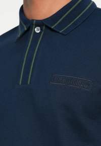 Camisa polo azul-marinho com malha texturizada, riscas verdes em contraste no colarinho e logótipo em relevo no lado esquerdo do peito.