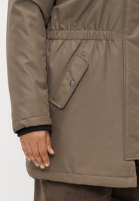 Casaco isolante castanho com cintura ajustada, apresentando um bolso lateral com fecho de botão e punhos canelados. Textura de tecido suave.