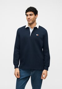 Άνδρας που φορά μπλε μακρυμάνικο ρούφαν ράγκμπι με λευκό γιακά και μικρό λογότυπο, συνδυασμένο με μπλε τζιν, στέκεται μπροστά σε απλό φόντο.