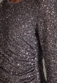 Top en sequins gris métallique, présentant une surface texturée et des détails froncés sur les côtés. Le tissu reflète la lumière avec un aspect brillant.