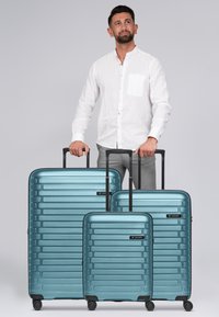 Tres maletas rígidas en teal metálico, con rayas horizontales y acentos en negro, colocadas junto a un hombre vestido con una camisa clara.