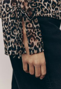Bluzica s uzorkom leoparda i prozirnom teksturom, koja sadrži resicu na rukavu učvršćenu mašnicama. Crni traper vidljiv ispod.