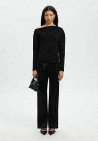 Czarna bluzka z długim rękawem i asymetrycznym dekoltem, w połączeniu z szerokimi czarnymi spodniami i czarnymi szpilkami, trzymająca małą czarną torebkę.